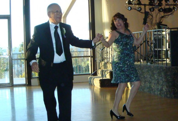 A man in a black suit dancing with a woman in a dark-print, knee-length dress and black heels. The are on the dance floor at a wedding reception.