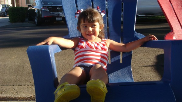 an almost 3-year-old girl in a red-and-white striped top and blue shorts sitting in a big, plastic blue chair and smiling.